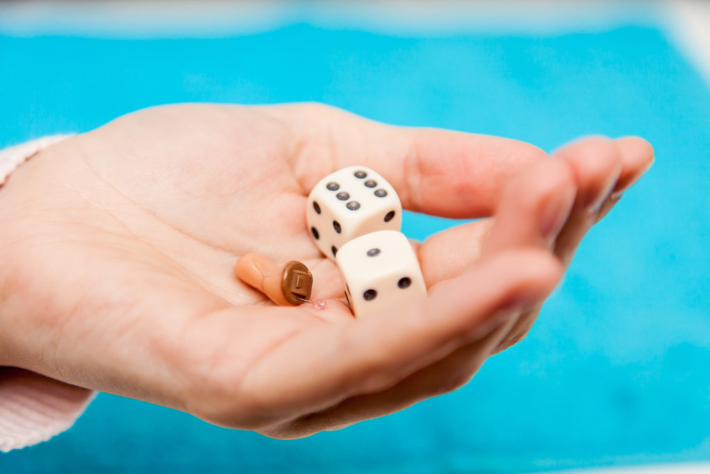 Zwei Würfel und ein braunes Hörgerät liegen in einer offenen Hand vor einem blauen Hintergrund, symbolisierend die Verbindung von traditionellem Spielvergnügen mit unterstützender Hörtechnologie